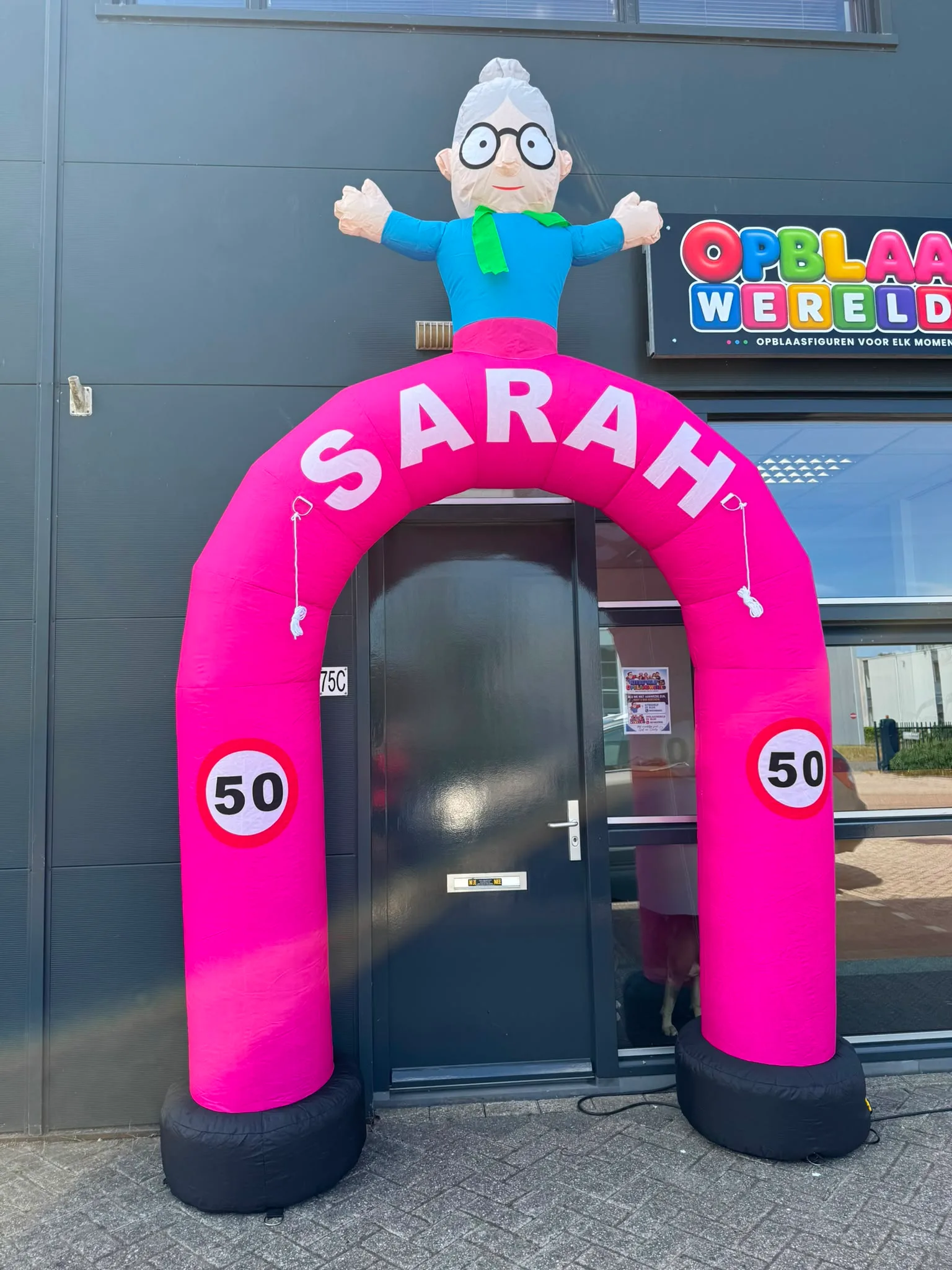 Roze opblaasboog van 3,5 meter met de tekst SARAH, een Sarah-figuur met bril en groene sjaal bovenop en twee borden met het getal 50, opgesteld voor de deur.