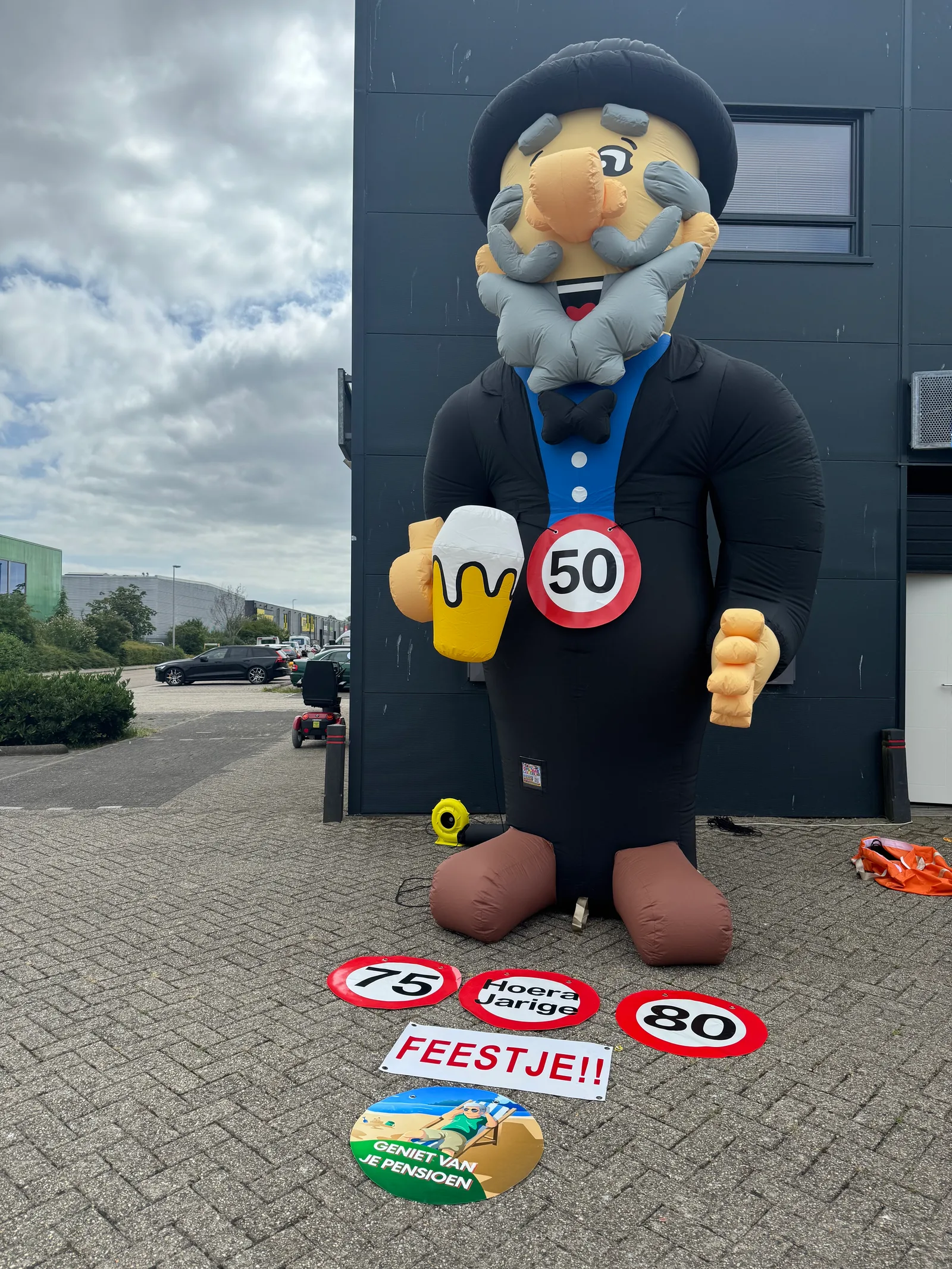 Grote opblaasfiguur van Abraham in zwart pak met hoed, grijze baard en bierpul, naast borden met 50, 75, 80 en Geniet van je pensioen, voor het pand van Opblaaswereld de Blok.
