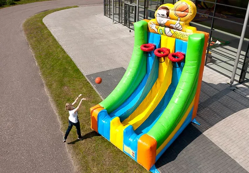 Opblaasbaar basketbalspel van 5,5 meter hoog in groen, geel, blauw en oranje met drie rode ringen. Een persoon gooit een oranje bal richting de baskets.