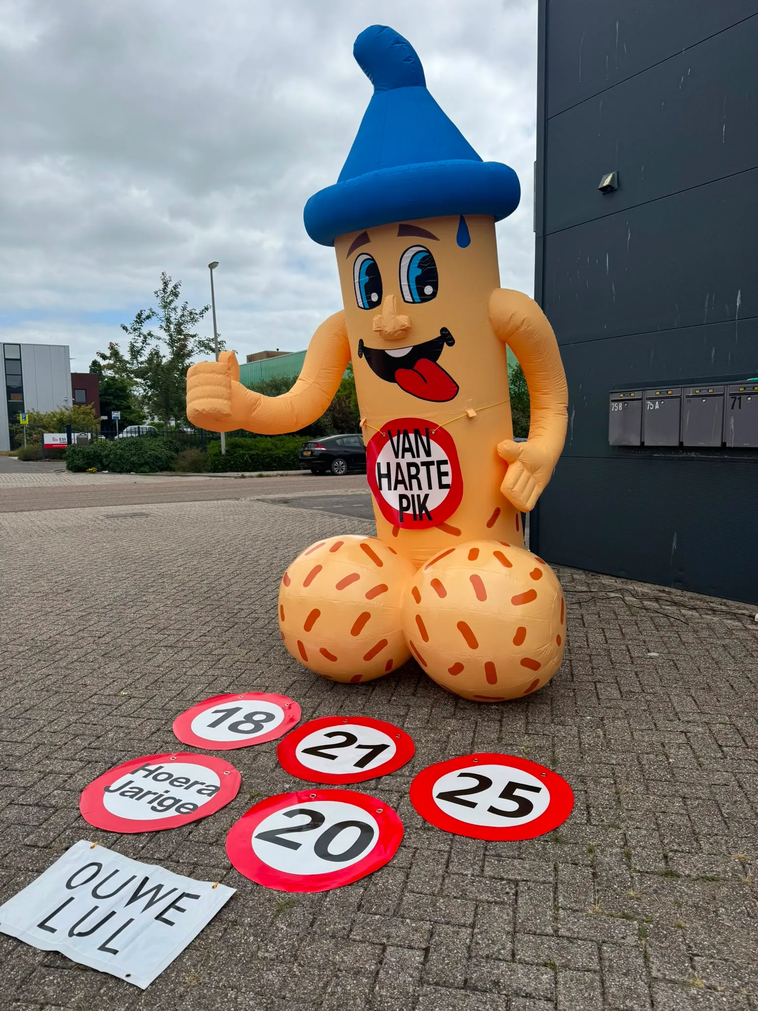 Opblaasbare Ouwe Lul van 3,5 meter met blauwe muts en banner met de tekst Ouwe Lul, met daarvoor leeftijdsborden 18, 20, 21 en 25, opgesteld voor het pand van Opblaaswereld de Blok.