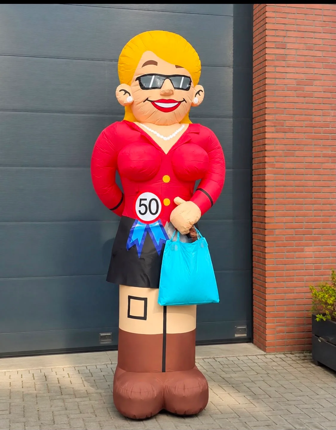 Opblaasbare Sarah feestpop van 3,5 meter in rood jasje met zonnebril en blauwe boodschappentas, naast een bord van Opblaaswereld de Blok en leeftijdsrosetten met 18, 25, 50 en 70 op de grond.