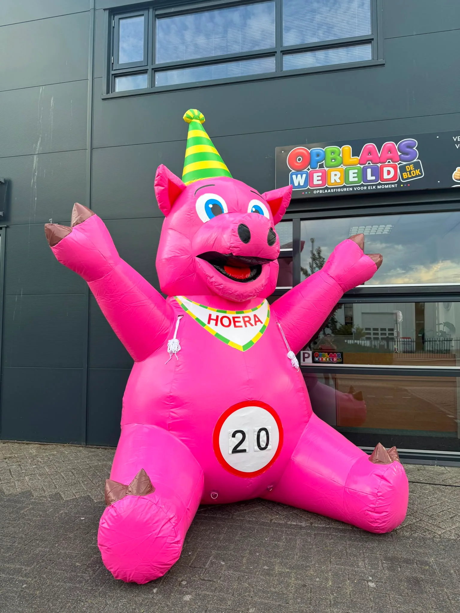 Roze opblaasbaar feestvarken van 3,5 meter met open armen, een 'Hoera' banner en een bord met het getal 40, opgesteld voor het pand van Opblaaswereld de Blok.