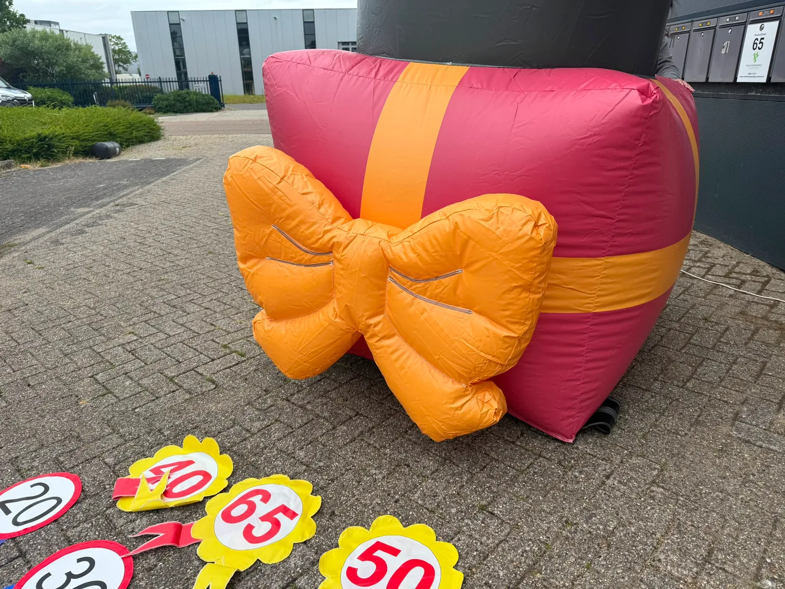 Opblaasbare Sarah pop van 4 meter met feesthoedje en rosette 40, staand op een roze cadeau met oranje strik, met rosetten en banner Feestje ervoor.