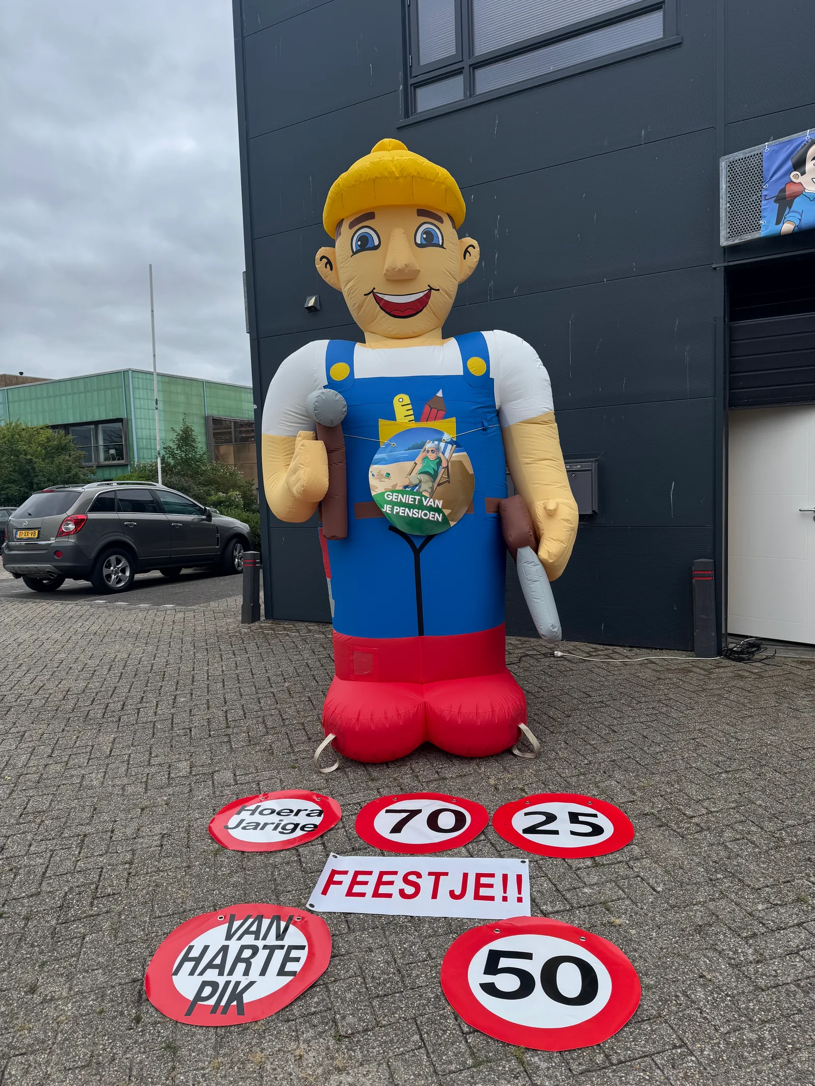 Opblaasbare Abraham feestpop van 3,5 meter als klusser met blauwe tuinbroek, hamer en spatel, met borden met teksten en leeftijden 70 en 50 ervoor.