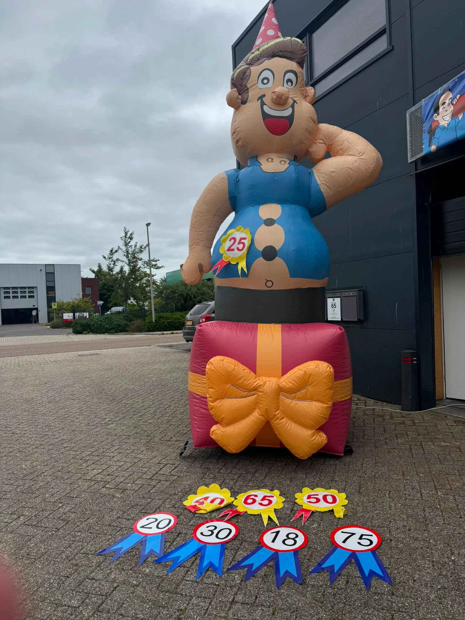 Opblaasbare Abraham van 4 meter met feesthoedje op een roze cadeau met gele strik, voor het pand van Opblaaswereld de Blok, met leeftijdsrozetten op de grond.