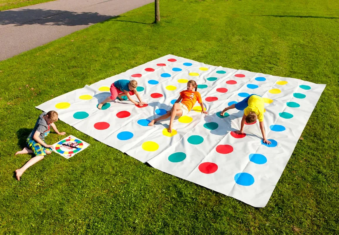 Grote witte Twister-mat van 5 bij 5 meter met gekleurde stippen op het gras, waarop vier kinderen spelen en een kind aan de draaischijf zit.