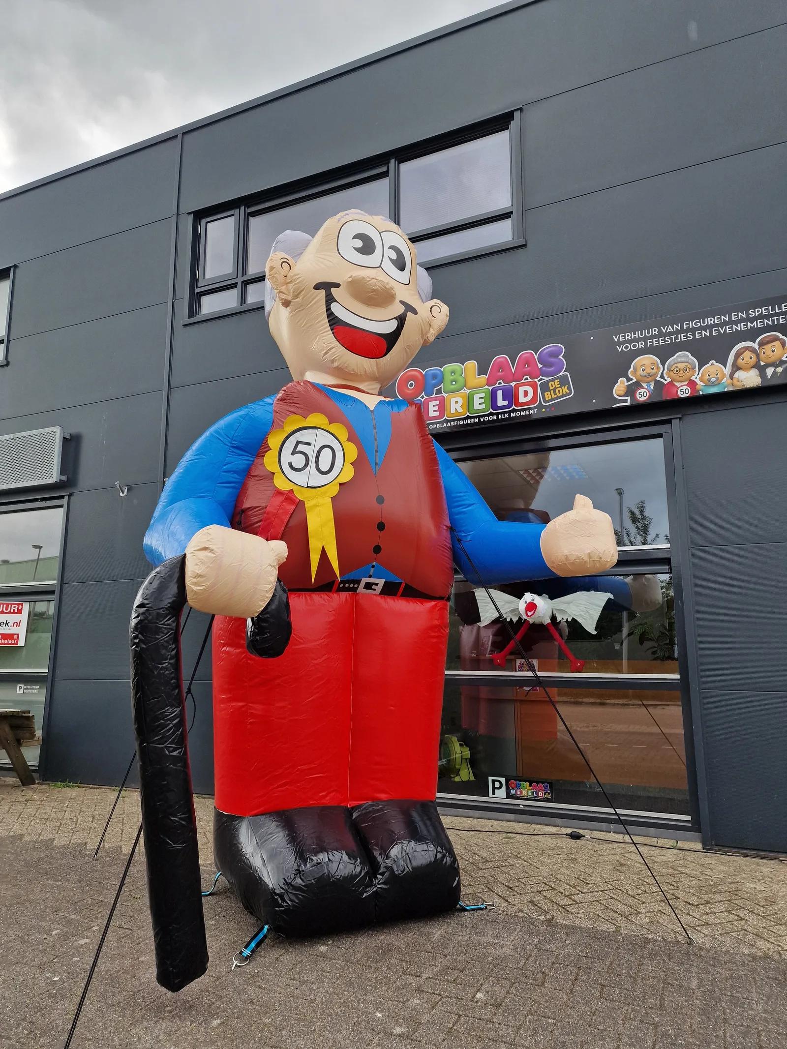 Opblaasbare feestpop Abraham van 4 meter hoog met grijs haar, rood vest, blauwe mouwen, een zwarte wandelstok en een rozet met het getal 50, voor het pand van Opblaaswereld de Blok.