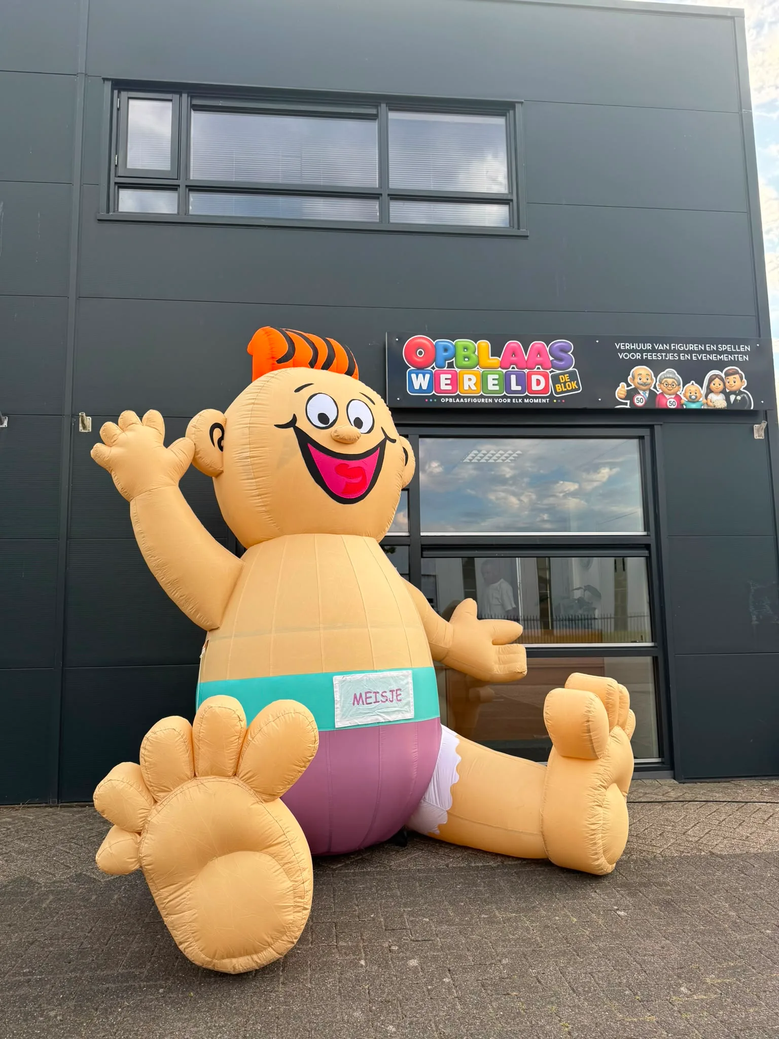 Opblaasbare baby van 3 meter met oranje kuif, zwaaiende hand en luier met label 'meisje', staand voor het pand van Opblaaswereld de Blok.