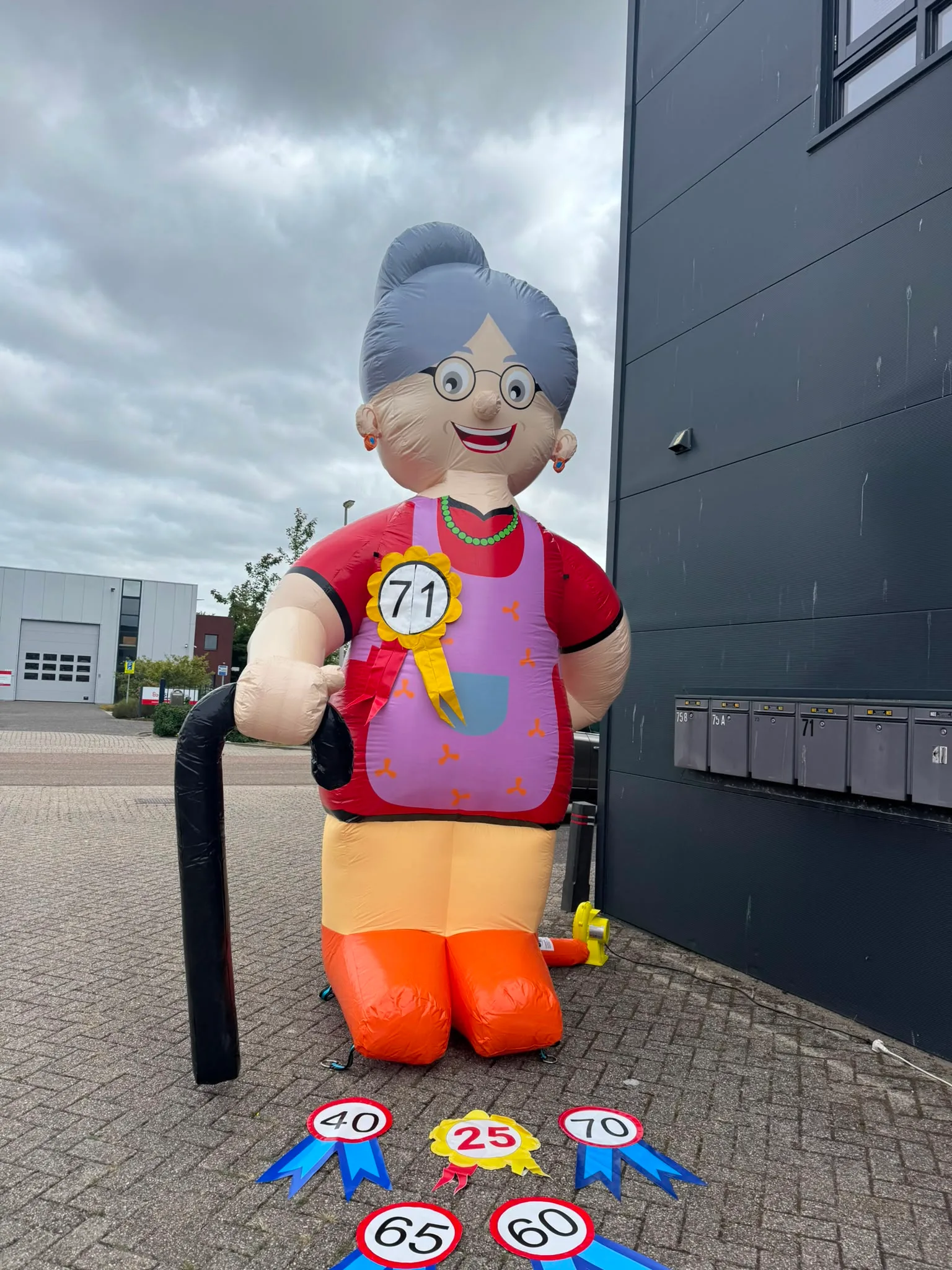 Opblaasbare Feestpop Sarah van 4 meter met grijs haar, bril, schort en stok, met leeftijdsrosetten op de grond voor het pand van Opblaaswereld de Blok.