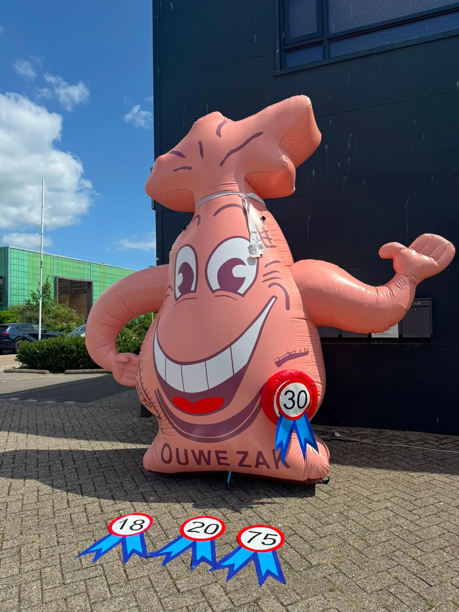 Opblaasbare Ouwe Zak van 3,5 meter in oranje met vrolijke lach, zwaaiende hand en verkeersbord 50, opgesteld voor het pand van Opblaaswereld de Blok.