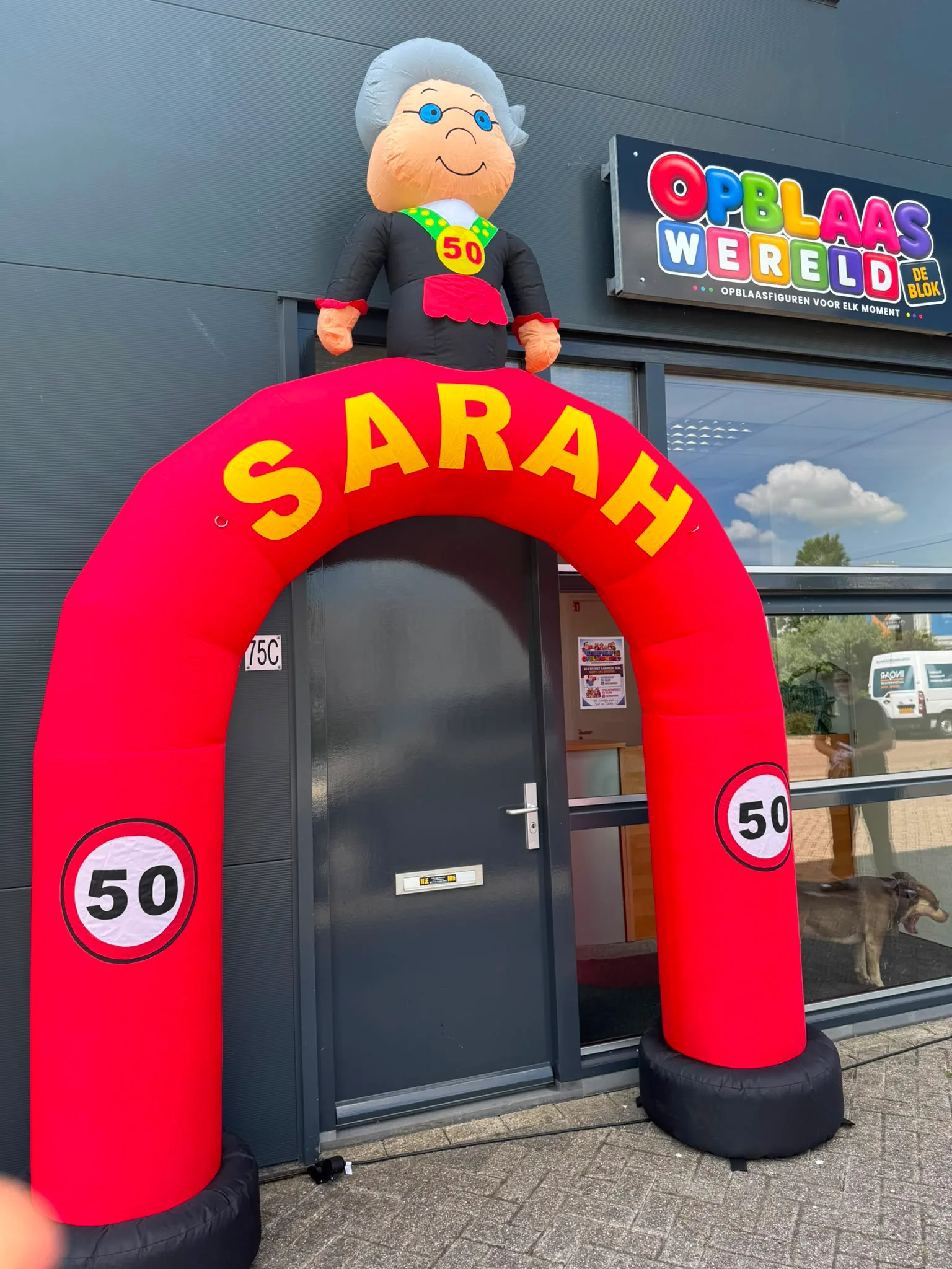 Rode opblaasboog met de tekst SARAH in gele letters, twee 50-borden en een Sarah-pop met bril bovenop, geplaatst voor de deur van Opblaaswereld de Blok.