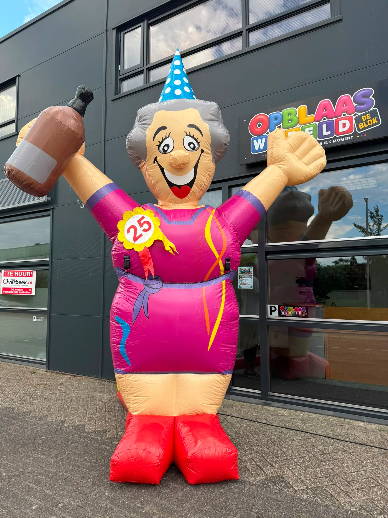 Opblaasbare Sarah Feestpop van 4 meter hoog in roze jurk met feesthoedje, fles in de hand en rozet met het getal 25, staand voor het pand van Opblaaswereld de Blok.